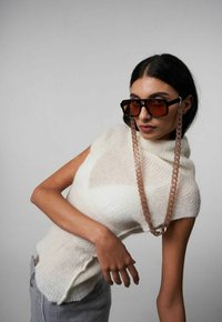 Top bianco lavorato a maglia senza maniche con una finitura texturizzata, abbinato a occhiali da sole oversized neri e a una catena trasparente per occhiali.