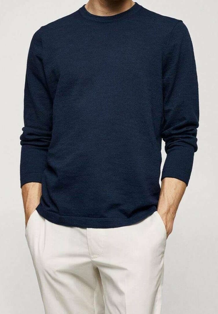 Homme portant une chemise bleu marine foncé à manches longues avec un pantalon couleur crème, debout les mains dans les poches devant un fond uni.