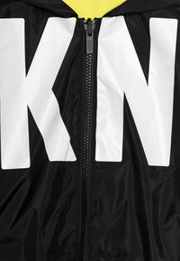 Musta tuulitakki, jossa on valkoinen "KN"-logo, vetoketjullinen etuosa ja keltainen hupun vuori lisäkontrastia ja yksityiskohtia varten. Sileä pinta.