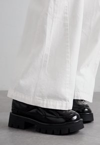 Black chunky platform shoes with a glossy finish and textured leather accents, worn with white wide-leg pants, highlighting contrasting colors.