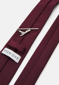 Cravat bordo realizată dintr-o țesătură netedă, cu un clips de cravată argintiu și un etichetă cu inscripția "PIER.ONE". Cravata are pliere și cusături ordonate.