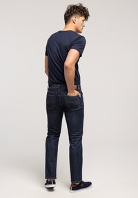 Młody mężczyzna z kręconymi włosami, ubrany w granatowy t-shirt, ciemnoniebieskie jeansy i granatowe trampki, stojący z jedną ręką w tylnej kieszeni.