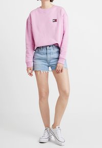 Rosa sweatshirt med en liten märkeslogotyp, matchat med ljusblå denimshorts med fransade kanter. Vita sneakers fulländar outfiten.