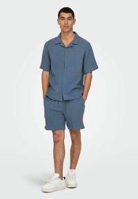 Lichtblauw T-shirt met korte mouwen en een subtiele vierkante textuur, gecombineerd met bijpassende shorts. Model draagt witte sneakers. Knopenlijst aan de voorkant.