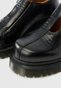 Μαύρα δερμάτινα μποτάκια μέχρι τον αστράγαλο με υφή κροκοδείλου, στρογγυλεμένη μύτη και χοντρές ραβδωτές σόλες. Διαθέτουν διακοσμητικές εγκοπές κατά μήκος των πλευρών.