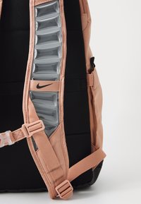 Mochila de Nike con un exterior de color rosa suave, secciones acolchadas en negro, una correa texturizada y un panel transparente brillante a lo largo del costado.
