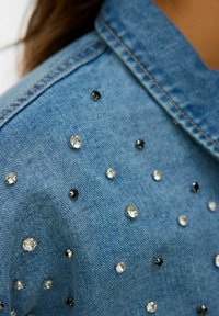 Giacca di jeans di colore azzurro chiaro, con strass trasparenti e neri distribuiti qua e là. Il tessuto ha un aspetto tessuto e testurizzato.