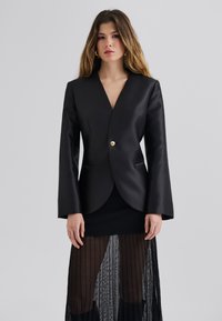 Blazer en simili cuir noir avec un décolleté en V profond, des manches longs évasées et un bouton doré unique. Superposé sur une jupe noire transparente et texturée.