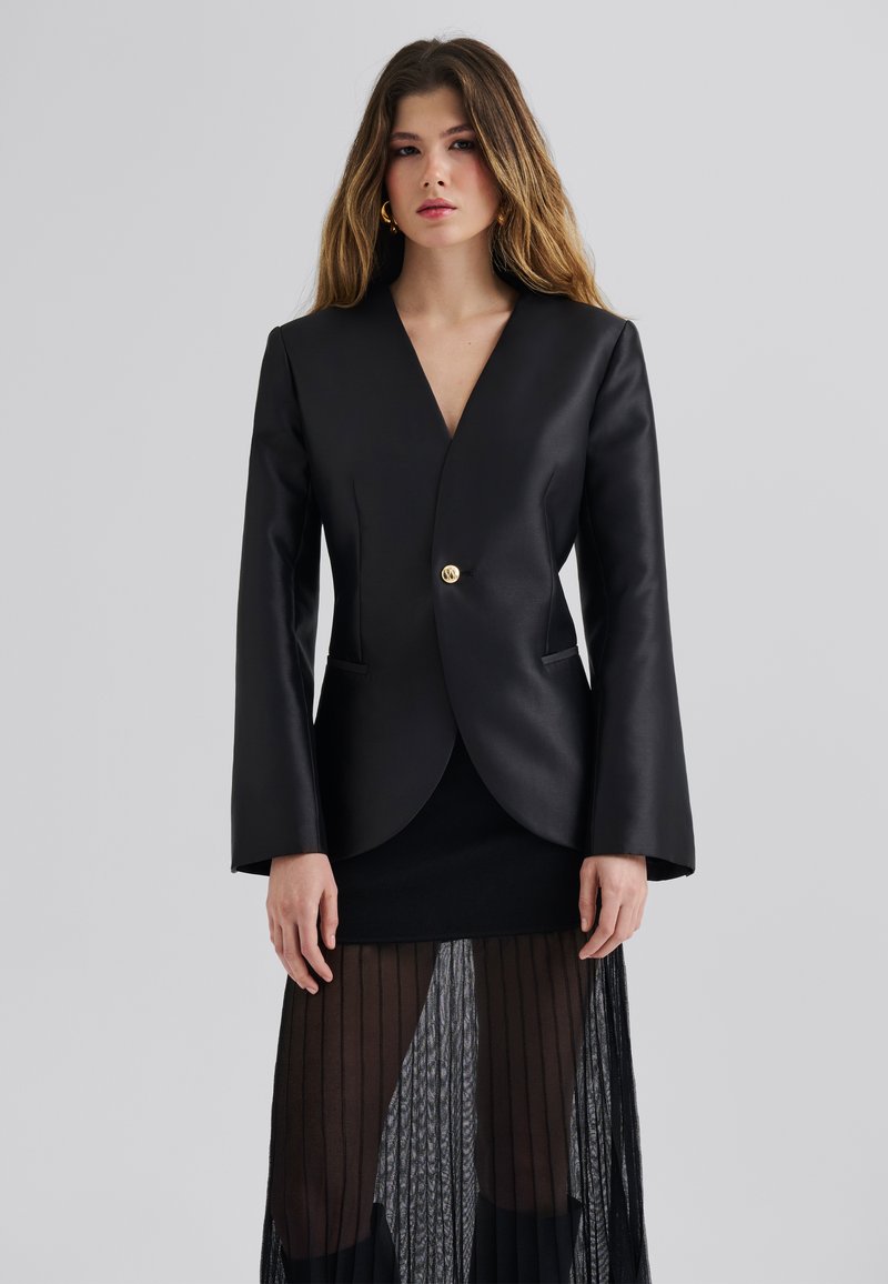 Blazer en simili cuir noir avec un décolleté en V profond, des manches longs évasées et un bouton doré unique. Superposé sur une jupe noire transparente et texturée.