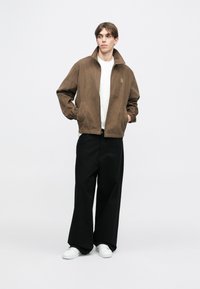 Brun cordfløyelsjakke med høy krage, elastiske mansjetter og frontglidelås, båret over en hvit skjorte og kombinert med svarte vide bukser.