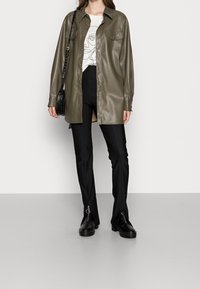 Cappotto camicia in ecopelle verde oliva con bottoni a pressione, abbinato a una t-shirt bianca con grafica e pantaloni neri a zampa con cerniere. Scarpe nere.