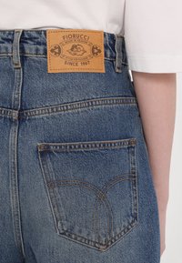 Srajčne jeans z visokim pasom, modre barve, z usnjeno oznako z blagovno znamko "Fiorucci" in značilnim oblikovanjem šivov na žepih.