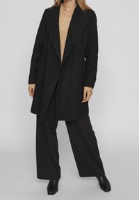 Musta villa-sekoite takki, jossa on avoin etusuunnittelu, lovikaulus ja pitkät hihat. Puettua tummanruskean teksturoituneen neuleen ja leveiden lahkeiden housujen päällä.