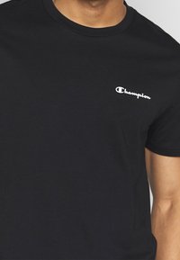 Sort bomulds t-shirt med korte ærmer og rund hals. Har et hvidt "Champion" logo på venstre bryst. Glat tekstur, standard pasform.