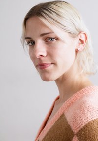 Pulover din lână cu decolteu în V, având un model în carouri în nuanțe moi de piersici și maroniu. Păr scurt, blond deschis, cercei simpli tip stud.