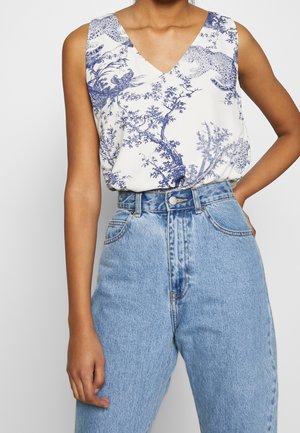 Haut blanc sans manches à col en V avec imprimé bleu de nature et d'animaux, rentré dans un jean en denim bleu clair taille haute, main sur la hanche.