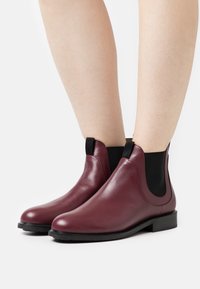 Botas de tobillo de cuero burdeos con paneles laterales elásticos, bajo tacón negro y textura suave, con un diseño elegante y minimalista.