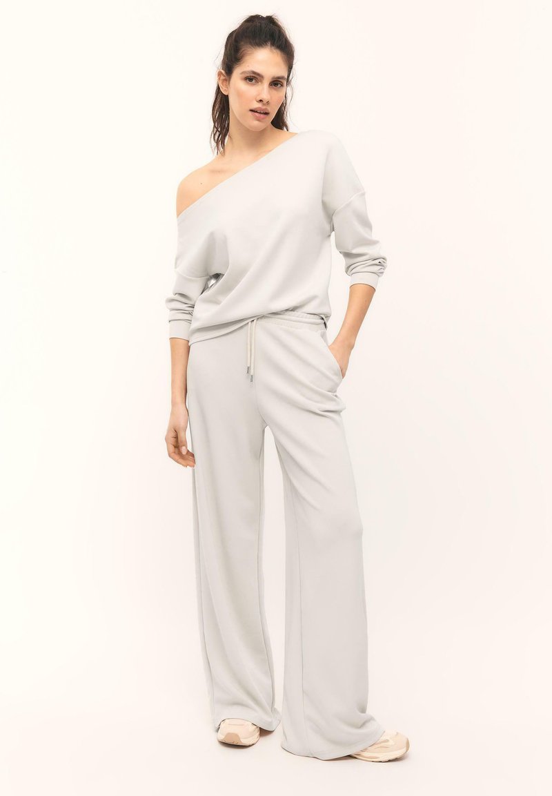 Jeune femme portant un sweat-shirt gris clair à épaules dénudées et un pantalon large assorti avec des baskets, debout devant un fond uni.