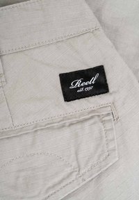 Hellbeige Stoff mit einer dezenten gestreiften Textur, versehen mit einer genähten Tasche und einem schwarzen Etikett, auf dem "Reell est. 1997." steht.