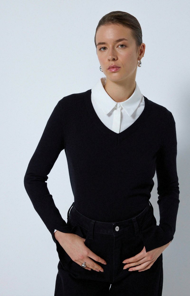 Pull marin à col en V, manches longues, superposé sur une chemise blanche à col. Un pantalon sombre et des bijoux simples complètent la tenue.