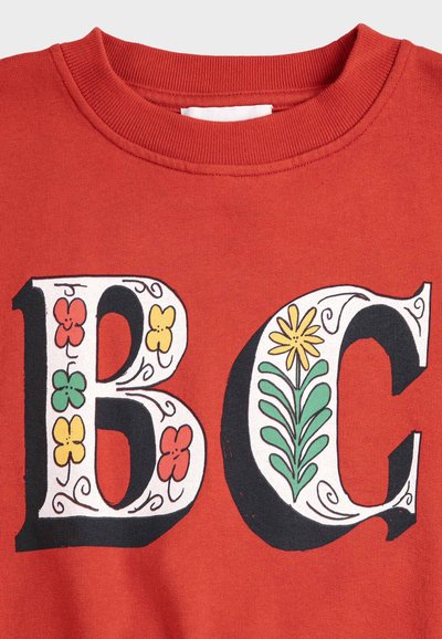 Tessuto rosso con grandi lettere decorative "B" e "C", ciascuna decorata con motivi floreali colorati e foglie verdi all'interno delle lettere.
