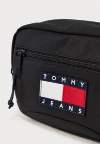 Nærbillede af sort stof-pose med frontlynlås og Tommy Jeans-logo i rød, hvid og marineblå.