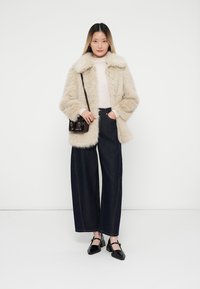 Cappotto in pelliccia sintetica beige con un ampio colletto, maglione crema sotto, jeans scuri di denim larghi e scarpe nere. Una piccola borsa marrone pende di lato.