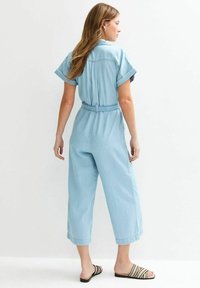 Combinaison en denim bleu clair avec des manches courtes, taille ceinturée et jambes raccourcies. Les sandales présentent des rayures noires et crème. Design simple et décontracté.