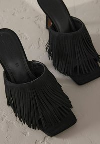 Mules en suède noir avec des détails en franges, présentant un design à bout carré et un talon modéré. Doublure intérieure lisse et semelles texturées.