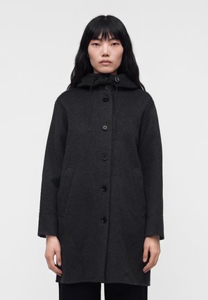Kvinde med langt sort hår iført en mørkegrå hættejakke med knapper foran og sidelommer, stående mod en ensfarvet baggrund.