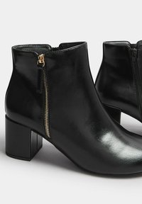 Botines negros brillantes con tacones cuadrados y detalles de cremallera dorada a un lado, presentados sobre un fondo blanco.