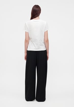 Femme aux longs cheveux bruns portant un t-shirt blanc uni à manches courtes et un pantalon noir à jambes larges, debout de dos sur un fond blanc.