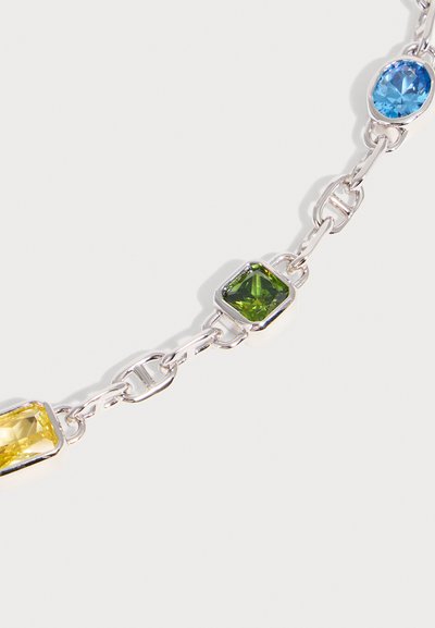 Bracelet chaîne en argent présentant des pierres précieuses rectangulaires et carrées bleues, vertes et jaunes montées dans des montures métalliques sur un fond clair.