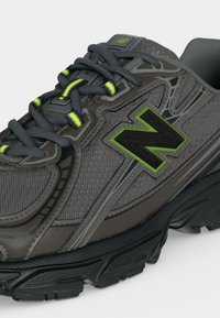 Σκούρο γκρι και μαύρο αθλητικό παπούτσι New Balance με υφασμάτινο δίχτυ, νεον πράσινες λεπτομέρειες και μαύρα κορδόνια με νεον πράσινους βρόχους σε λευκό φόντο.