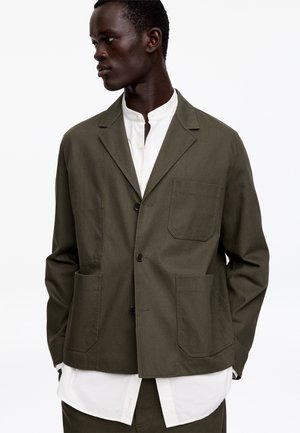 Homme en blazer décontracté vert olive avec trois poches avant, par-dessus une chemise blanche à col mandarin, regardant vers la gauche sur un fond uni.