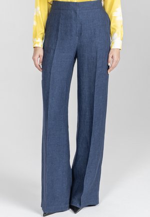 Kobieta ubrana w niebieskie, szerokie spodnie z wysokim stanem, zestawione z żółtą, długorękawową bluzką, stojąca na jednolitym tle.