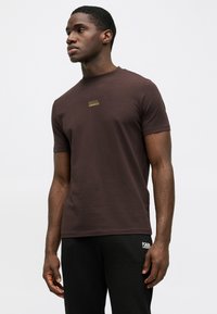 Tumši brūns īsās piedurknes T-krekls no mīksta auduma, ar mazu zelta logo uz krūtīm. Saskaņots ar melnām biksēm.