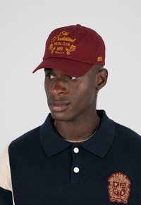 Bordeaux rote Mütze mit gesticktem goldenen Text und Grafiken, ausgestattet mit einem strukturierten Schirm. Getragen mit einem marineblauen Poloshirt und dekorativem Emblem.