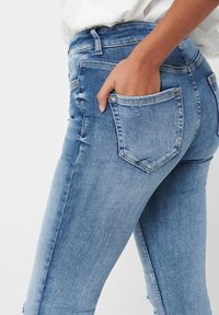 Person med ljusblå denimjeans och hand i bakfickan, mot en vit bakgrund.