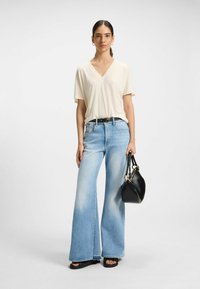 Beżowa bluzka V-neck, jasnoniebieskie dżinsy z szerokimi nogawkami, czarny pasek, czarna torebka z złotymi detalami, płaskie czarne sandały, neutralne tło.