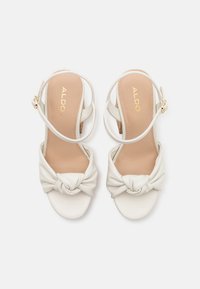 Sandales à talons blancs avec un design à l'avant noué, texture en cuir souple et fines lanières. Équipées d'une semelle intérieure beige légèrement rembourrée.