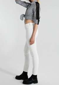 Jeans blancos ajustados con costuras laterales, combinados con una camisa de rayas en blanco y negro con un diseño corto y detalles de encaje, usados con botas negras de tobillo.