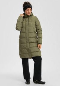 Žena nosi dug zeleni pufasti kaput, crne hlače, crne čizme i crnu traku za glavu, pozira s jednom rukom blizu ovratnika kaputa.