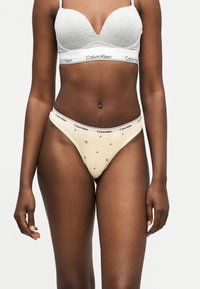 Γυναίκα που φοράει ελαφρώς γκρι σουτιέν Calvin Klein και μπεζ εσώρουχο Calvin Klein με μοτίβα μαύρου αστεριού και λογότυπου, στέκεται μπροστά σε απλό φόντο.