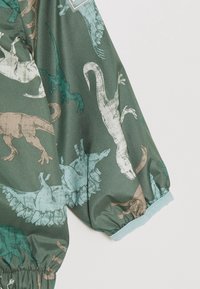 Grüner Stoff mit Dinosaurier-Drucken in Beige, Teal und Weiß. Elastische Bündchen und Saum; glatte Textur; semi-matte Oberfläche.