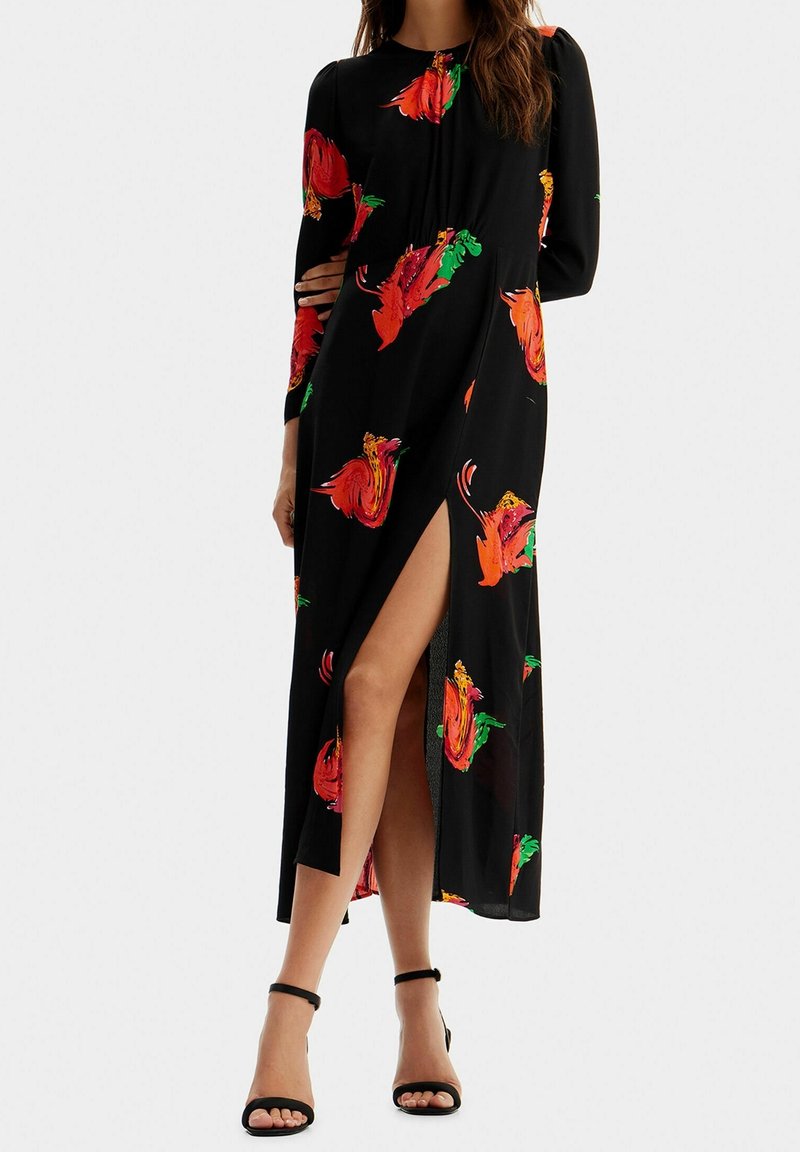 Robe noire à manches longues, ornée de motifs floraux vibrants en rouge, orange et vert. Une fente sur le côté et des sandales à bride de cheville sont visibles.