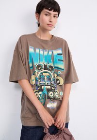 Brun oversized Nike T-shirt med et farverigt vintage grafik af højttalere og en cykel. Korte ærmer og rund hals. Casual stofstruktur.