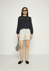 Mulher vestindo blusa preta, calções beiges, sapatos pretos e óculos de sol, segurando uma pequena bolsa com padrão de zebra contra um fundo simples.
