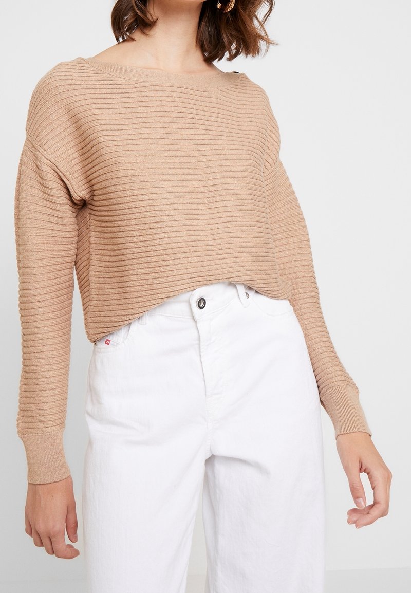 Lysebeige ribstrikket sweater med bredt halssnit og lange ærmer, kombineret med højtaljede hvide bukser. Tekstureret stofdetalje synlig.