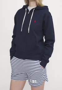 Kvinna som bär en marinblå zip-up hoodie med vita snören och randiga blå och vita shorts med en liten broderad logotyp på benet.